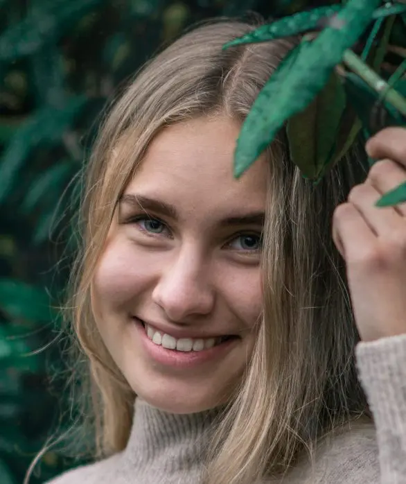 Dame med blondt hår som står inntil en vegg av grønne blader.