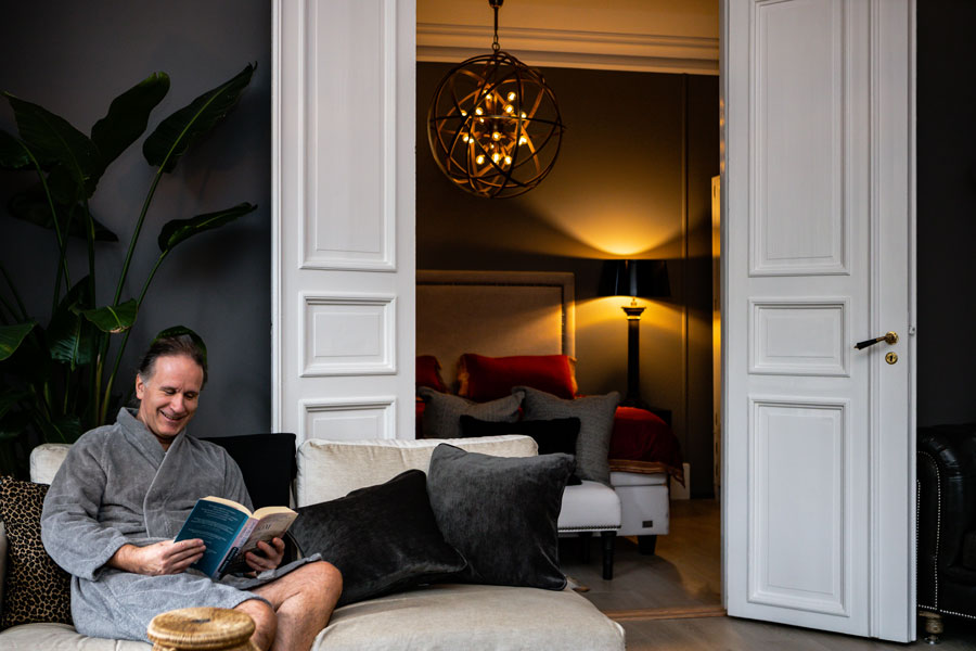 En smilende mann som leser bok på en sofa. Han er innført i en grå morgenkåpe i luksuriøst suite i klinkken.